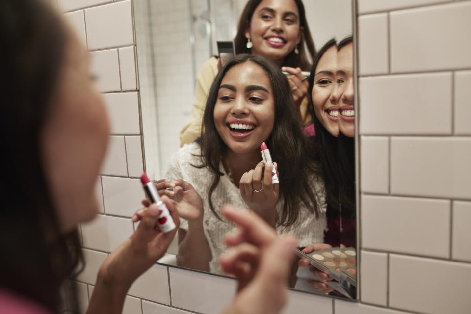 Three friends smile and apply makeup in front of a mirror